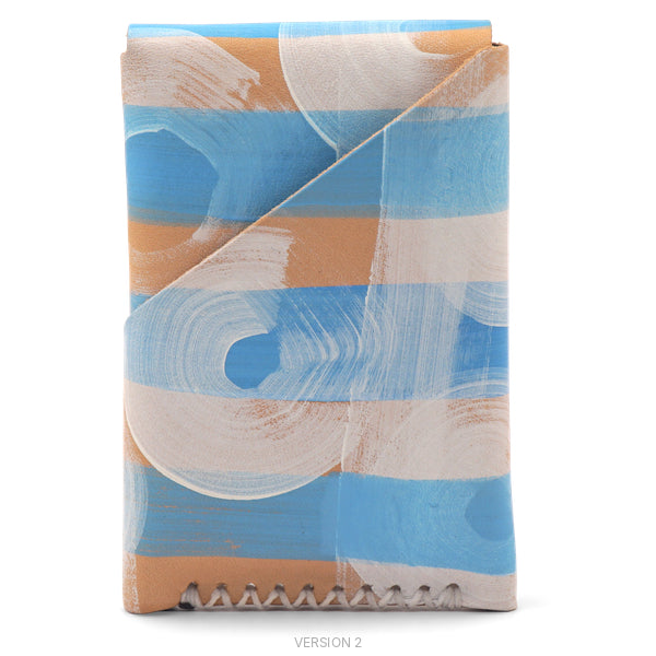 A folded clutch or wallet-style handbag with an abstract painted pattern in light blue, warm tan, and off-white, shown on a plain white background. The item has a flap closure and visible stitching along the bottom edge.