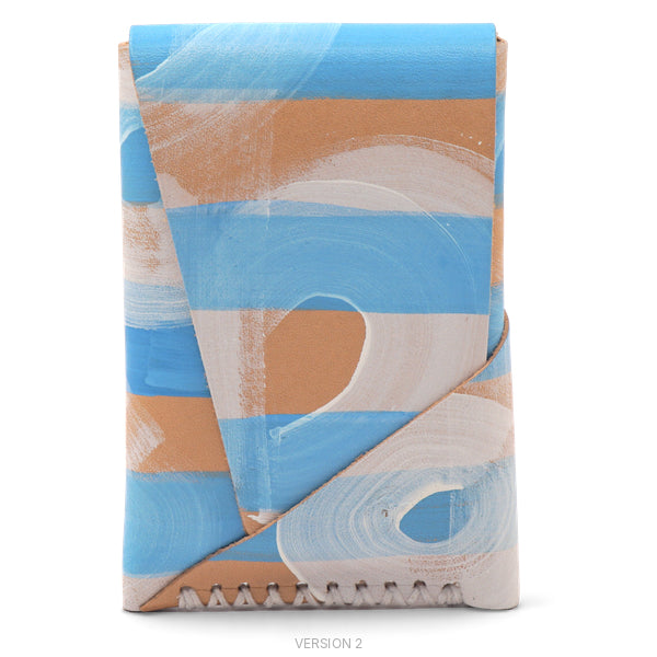 A folded leather wallet or card holder featuring a modern abstract pattern of horizontal stripes in light blue, natural veg tan leather, and textured white brush strokes, with a zigzag stitched edge at the bottom.